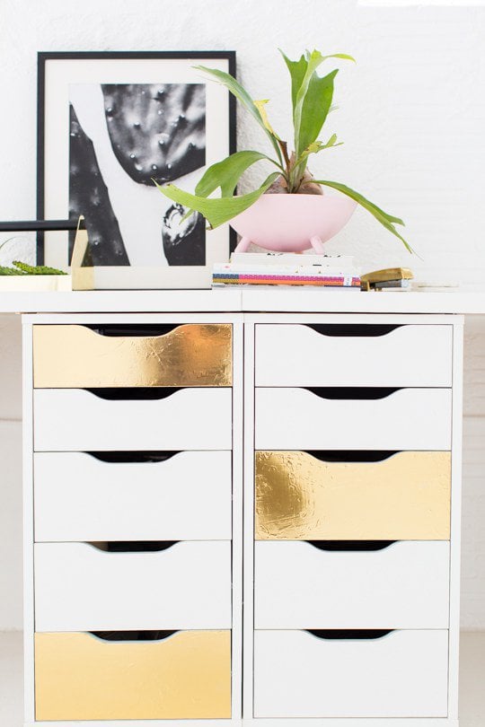 Two gold-foiled IKEA Alex drawers are next to each other under a white wooden DIY desk top with a plant and photograph on top. Altering drawers is one of the best DIY IKEA hacks. 