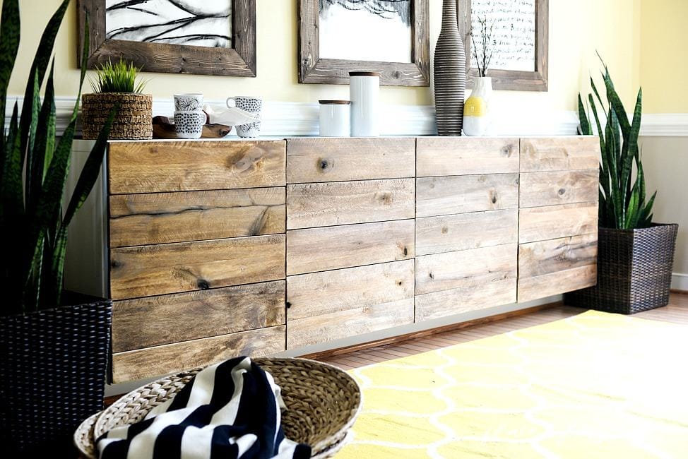 A long, reclaimed wood buffet table made from IKEA cabinets is against a pale-yellow wall decorated with picture frames and plants. 