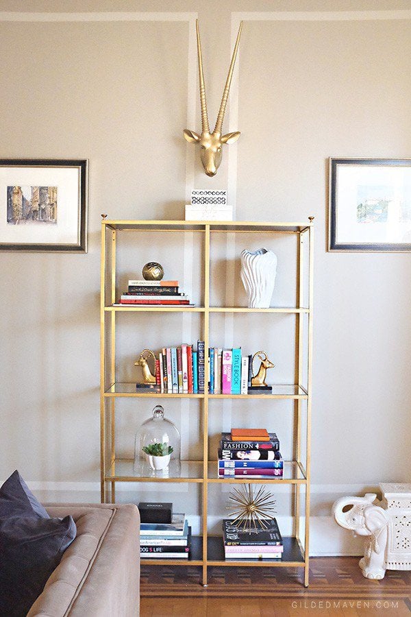 A gold storage rack made from IKEA Vittsjo holds books and other items. A gold horned animal head is above the open shelves, and framed pictures are on either side. 