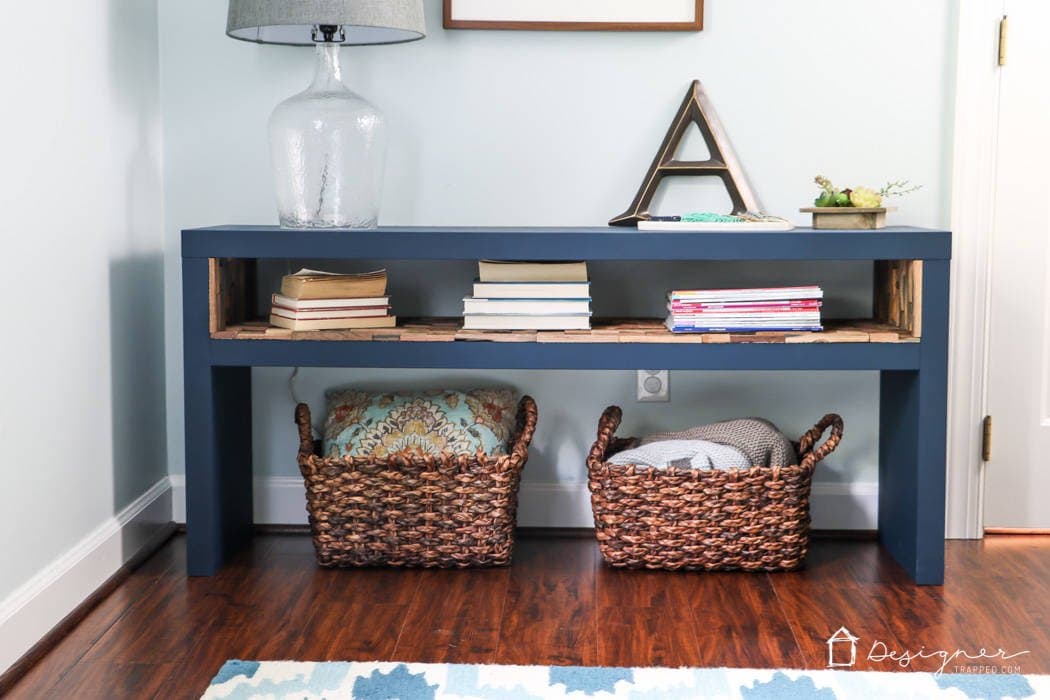 A blue sofa table is against a wall, with a wood-lined shelf, books, baskets, and other knickknacks on it. DIY IKEA hacks include Lack tables.