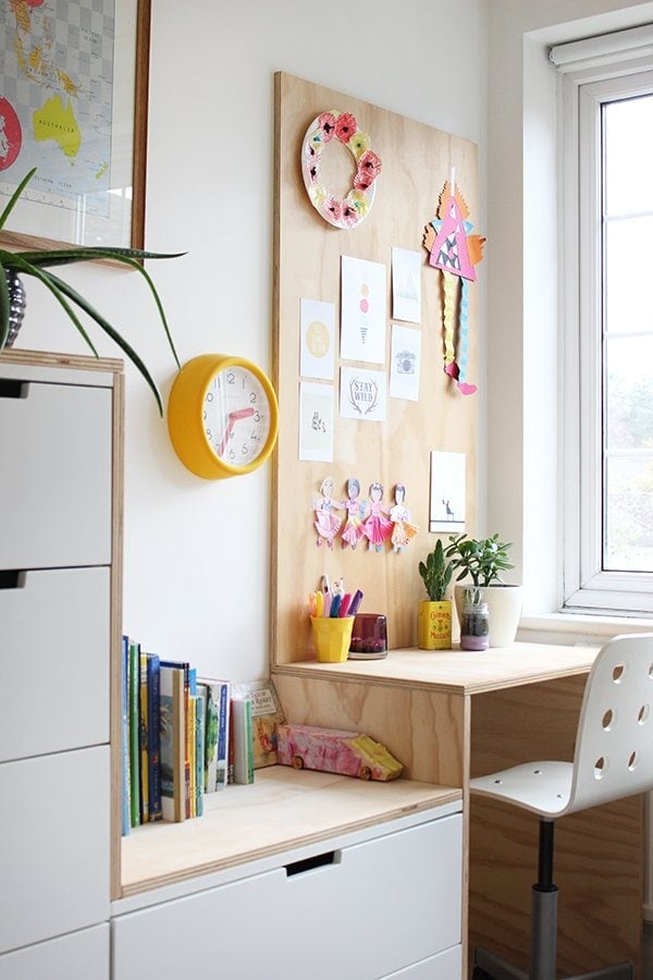 A compact plywood desk with a display of children's artwork on the wall, beside sizable IKEA storage bins that contain children's books and various supplies for a playroom. IKEA desk hacks can be for kids and adults.