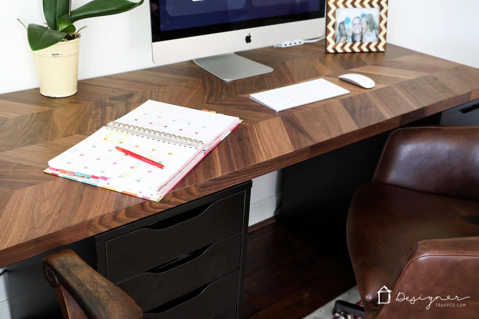 Part of a wooden desk top with an Apple monitor, calendar, and miscellaneous papers and photos showcases this DIY IKEA hack for office desk ideas. 