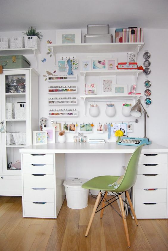 A crafting IKEA desk hack is made with all-white IKEA furniture, including drawers, a tabletop, shelving, and glass-door cabinets paired with a green chair. Craft supplies are arranged along the wall behind the desk.