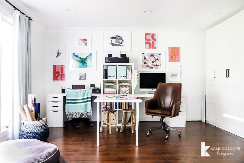 A double desk has a table between two work stations, a leather wheeled chair, drawers, shelving units and a gallery wall on a dark wood floor. IKEA desk hacks can be used for office and crafting space. The words "Kaleidoscope Loving.com" are in the corner. 