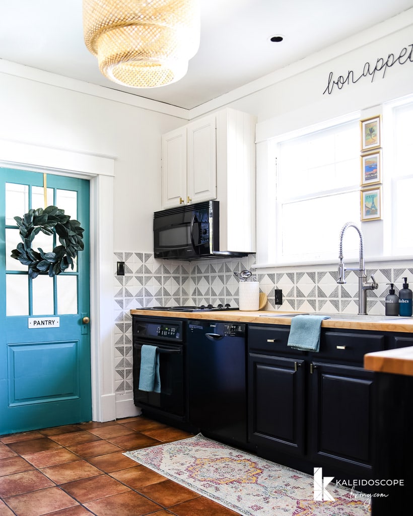 A kitchen corner with a blue door with a wreath and a sign that says "pantry," partial tiled walls, white upper cabinets and blackish lower cabinets.