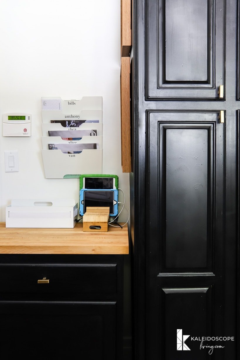 Part of a pantry door painted black, next to a wood countertop family command center with wall-mounted file trays with family member's names and a charging station for children's tablets.