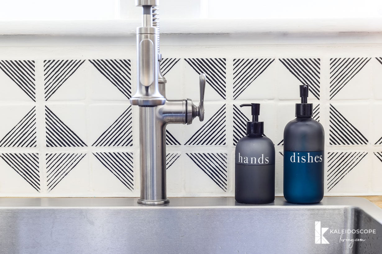 A close-up of a faucet and a white and black geographic tile stenciled backsplash. Two pump bottles to the right of the sink say "hands" and "dishes." "KaleidoscopeLiving.com" is written in the lower right corner.