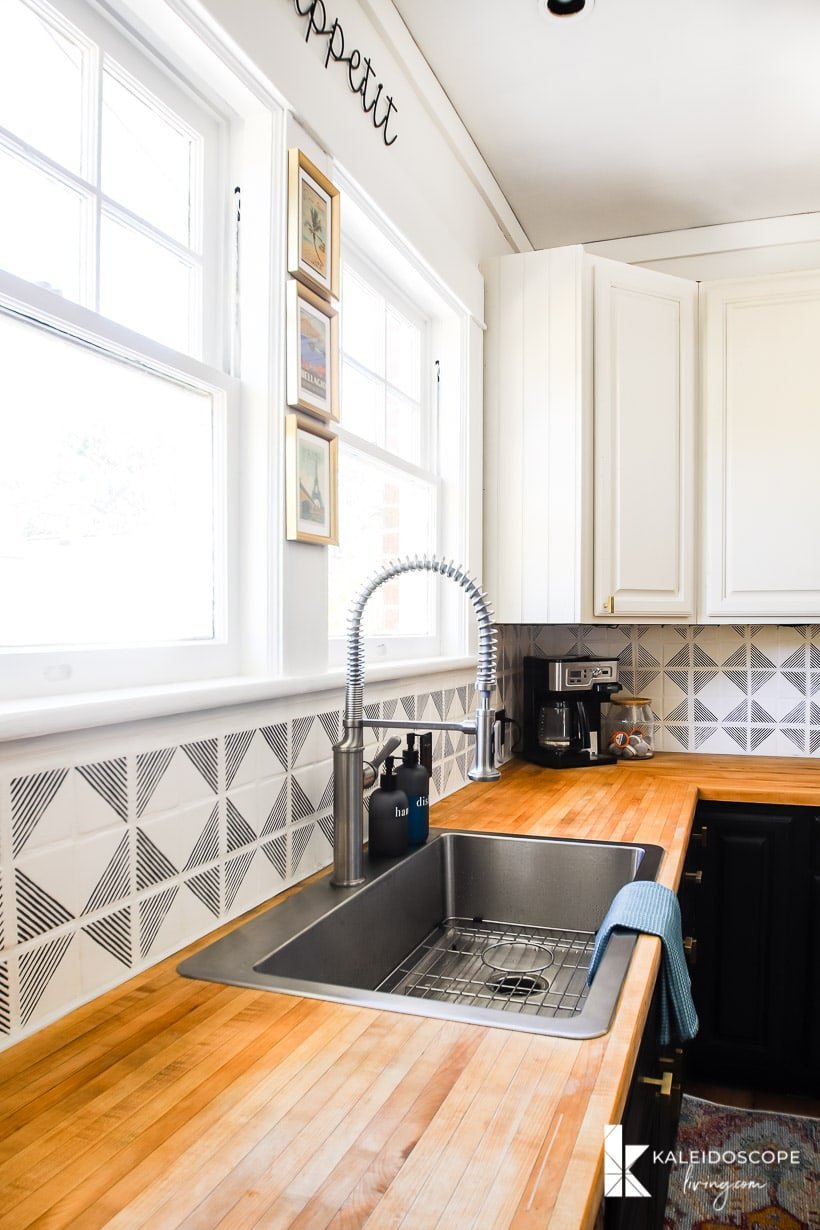 A black and white budget-friendly kitchen makeover has geometric black and white tiles as a backsplash, a treated wood countertop, stainless steel sink, two large windows and white cabinets. A coffee machine in the corner.