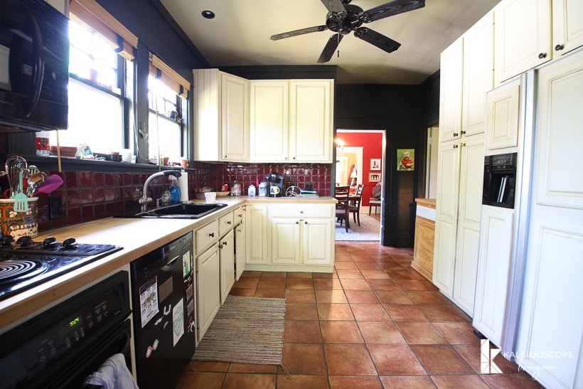 A view into a large but narrow outdated kitchen before budget-friendly kitchen makeover. The left side holds the counter, stove, oven, sink and cabinets and the right side has a white refrigerator, pantry and upper cabinets.