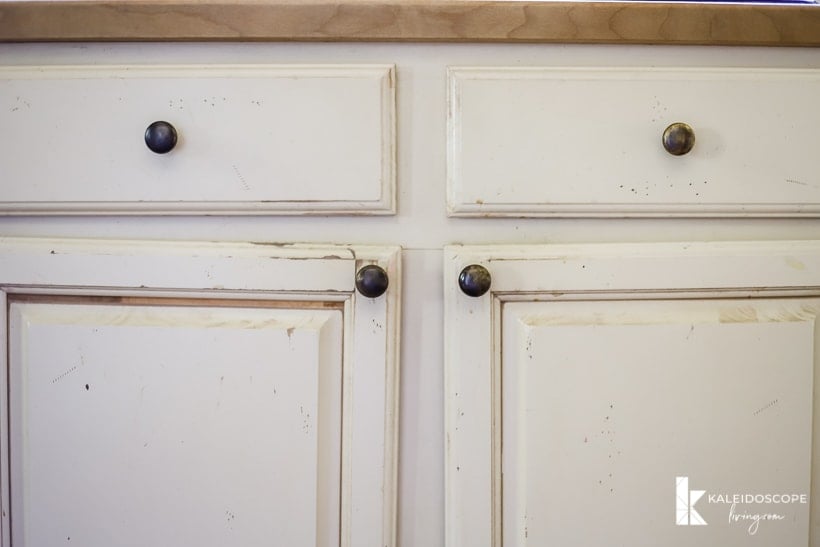 Worn out painted kitchen cabinets with uneven hanging doors, dirt, and paint scratches. The corner text reads, "KaleidoscopeLiving.com."