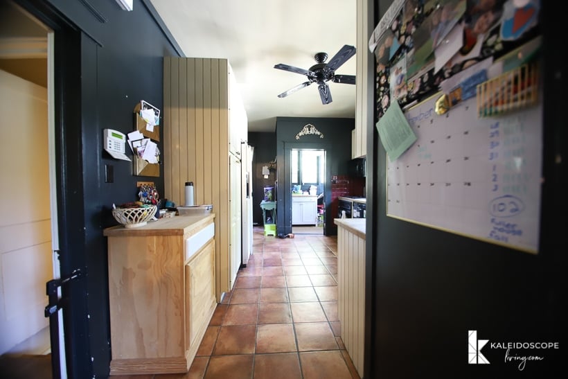 An outdated kitchen before a budget-friendly kitchen makeover removed the dark cave-like walls and trim. A family calendar hangs on the side of a refrigerator and brown tiles line the floor and hallway into the kitchen,