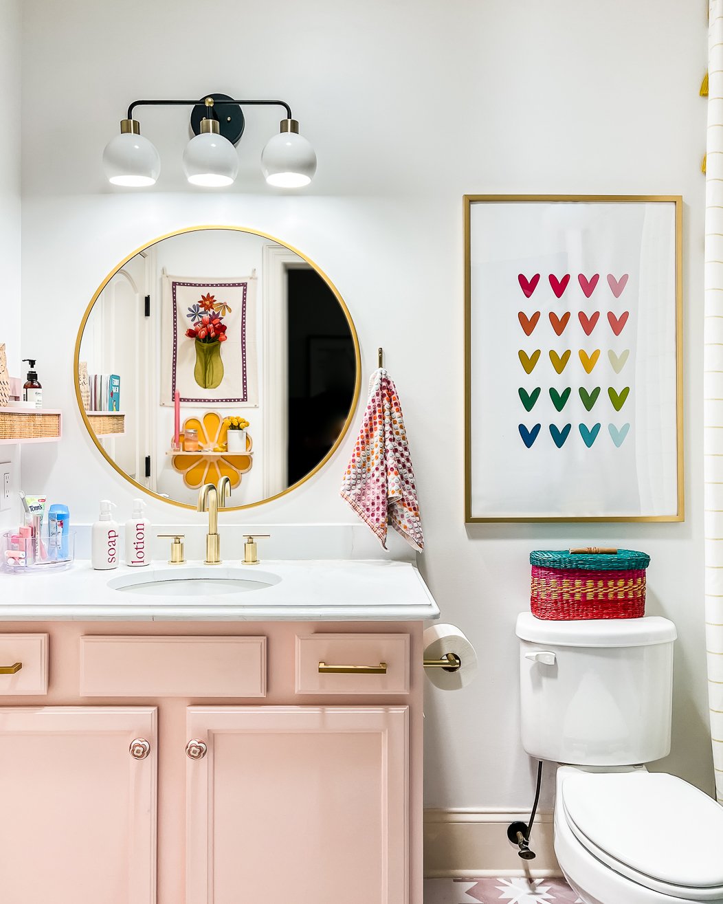 A bathroom makeover on a budget includes a round gold mirror, white walls with pink cabinets, rainbow heart artwork. toilet, and flower artwork reflected in the mirror. 