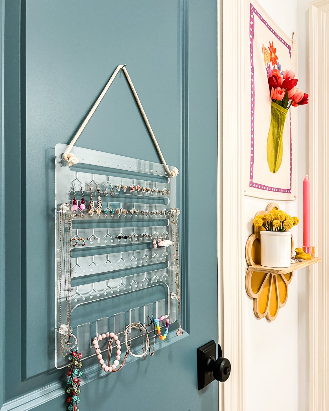 A clear acrylic hanging jewelry organizer in girl's bathroom on the back of a green-blue door with yellow flowers on the wall to the side. 