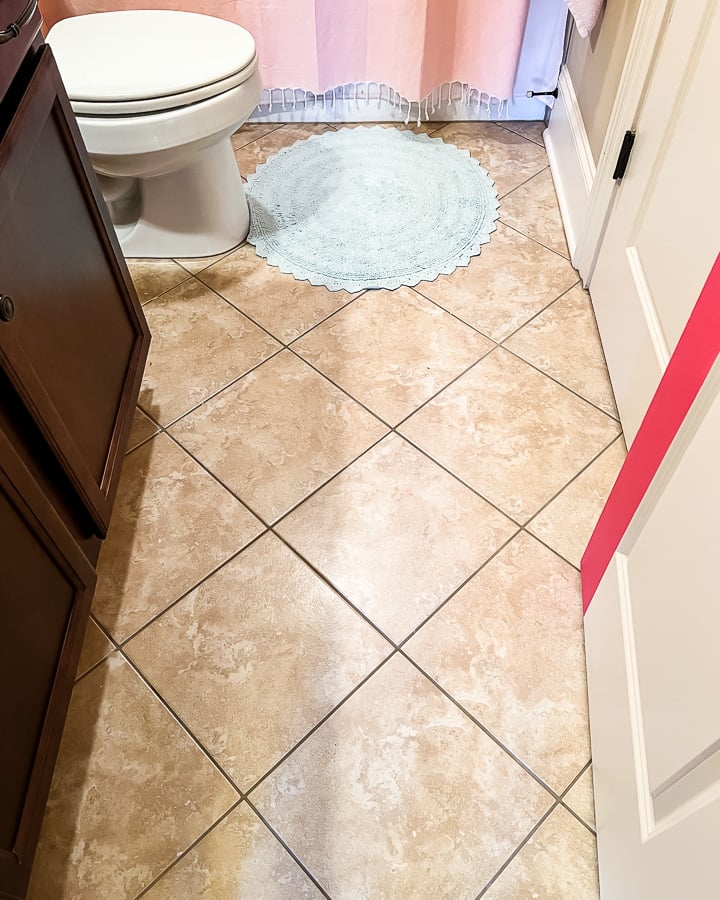 A before photo of an outdated brown bathroom floor tile with a dark brown cabinet and pink shower curtain. 