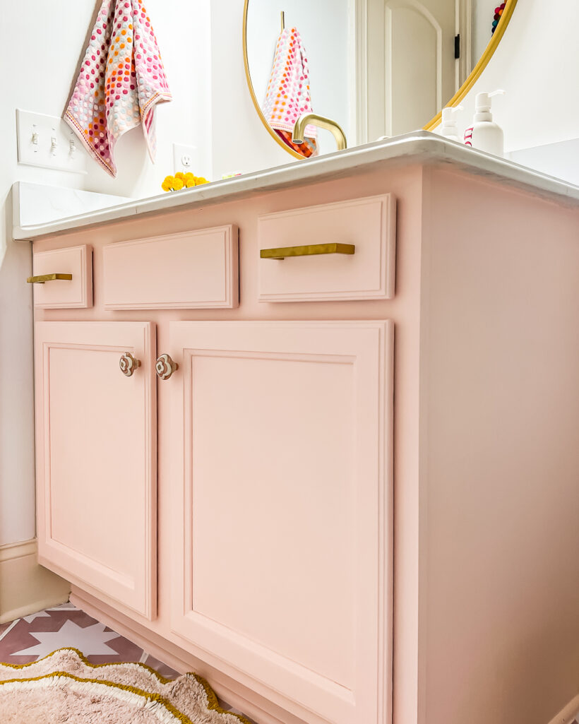 A light pink vanity in girl's bathroom, with gold hardware, a gold round mirror and white countertops. 