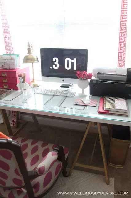 A stylish IKEA desk hack features a repurposed door placed on gold sawhorse legs, topped with glass, along with other office essentials such as a computer monitor, printer, and pink accessories. "www.dwellingsbydevore.com" is printed in the corner. 