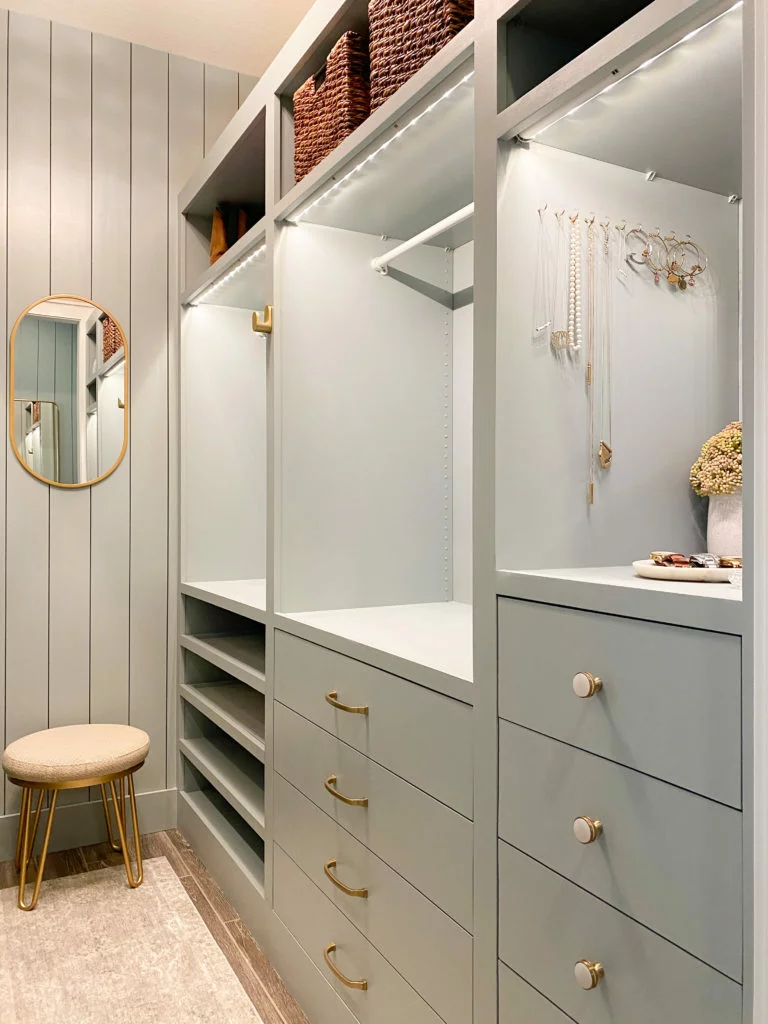 An angled view of customized sage green IKEA Pax units in closet with customized. Jewelry and wicker baskets are part of the storage configuration. A stool and mirror are in the back. 