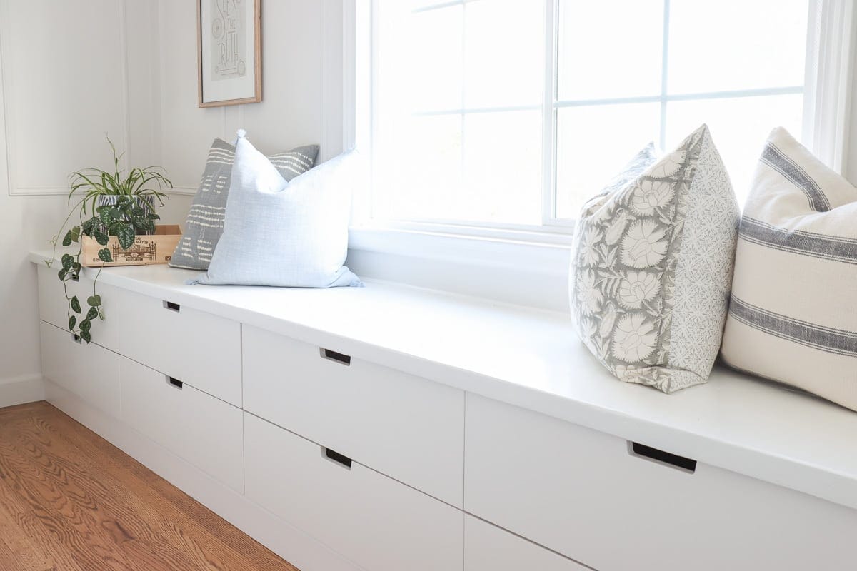 A white window seat made from IKEA Nordli drawers lines the living room wall under the window. The pillows and plant on the long bench have storage drawers below.