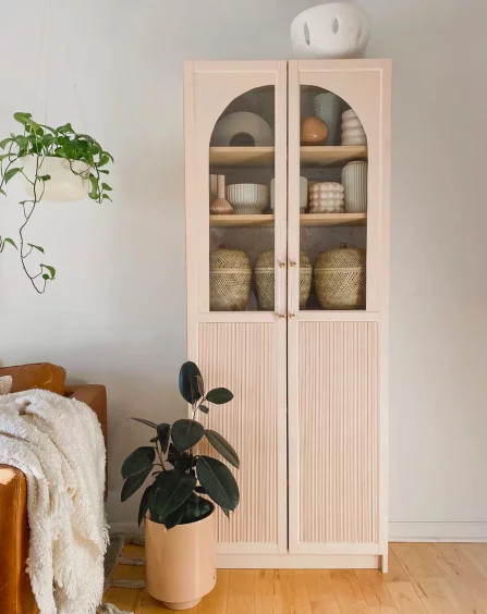 A tall cabinet with arched glass doors next to potted plants, a light leather chair and off-white blanket. This DIY IKEA hack combines Billy bookcases and IKEA Oxberg cabinet doors. 