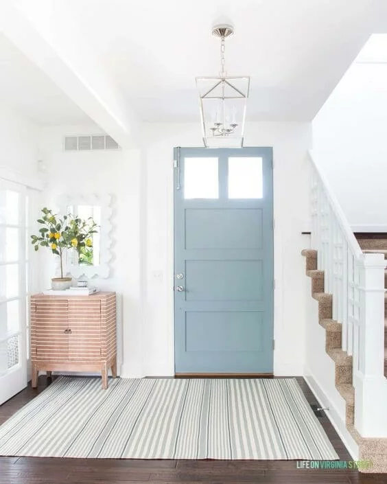 A light blue-gray front door next to a staircase and a side table showcases 