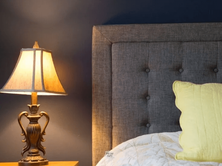 A closeup of a bedside lamp and  navy bedroom wall with upholstered gray headboard and yellow pillow 