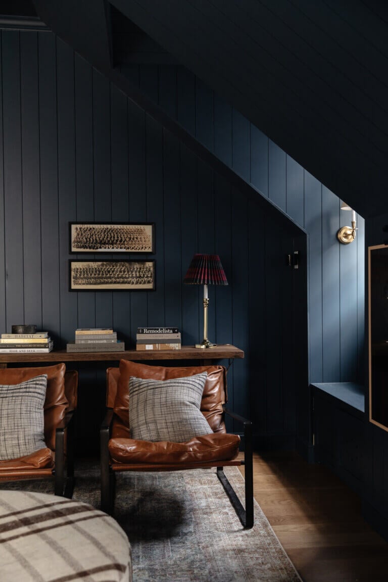 The corner of a bonus room with dark navy paneled walls. leather chairs and a tabletop with stacked books. 