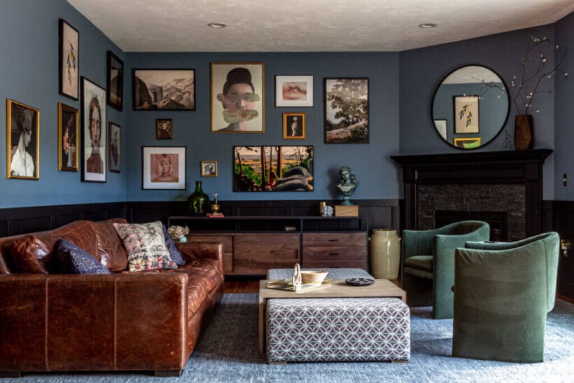 A moody living room with a blue gray paint color on the wall with gallery art and a leather couch. 