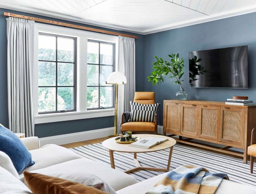 A bright family room has blue gray paint colored walls with light wood furniture and white curtains. 