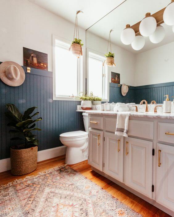 A corner of a bathroom with blue green wainscoting, white toilet and  counter, and a bright window. 