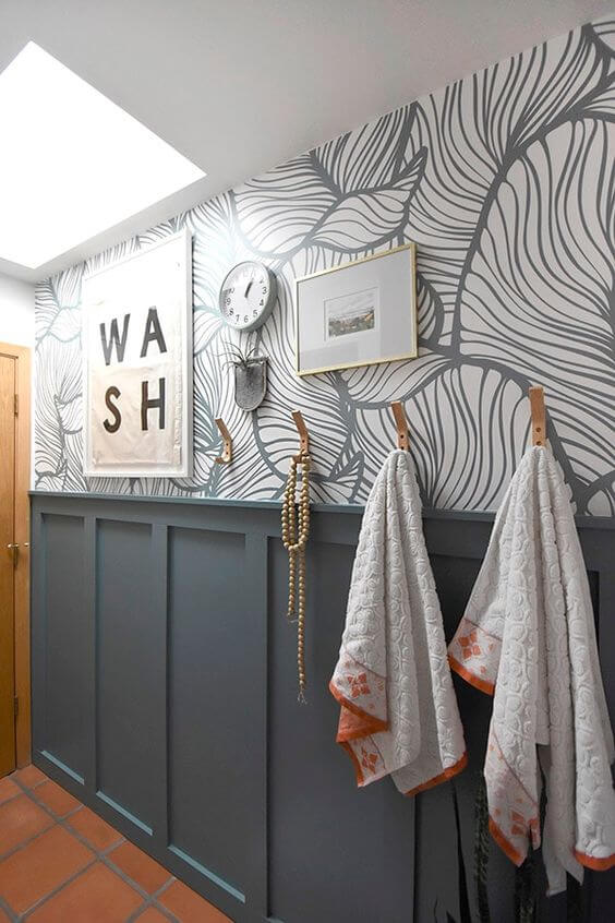 A bathroom wall with blue gray paint colors on wainscotting board and printed wallpaper. A sign says "wash" and towels hang on hooks. 