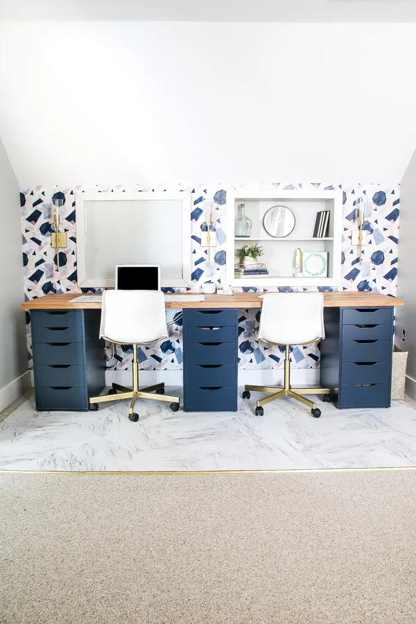 An IKEA desk hack featuring navy drawers, a natural wood countertop, and built-in shelves on a wall with blue-patterned wallpaper under an attic roof slope. 