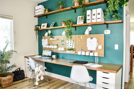 A homeschool area includes a long wooden tabletop with IKEA drawers underneath, two chairs, al facing a green wall with a peg-board and shelves for books and plants. The desk is extra long thanks to an IKEA desk hack. 