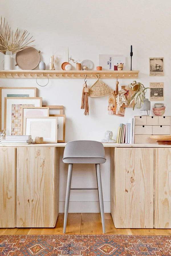 Neutral tones dominate this standing IKEA desk hack. A shelf with hooks hangs above a long desktop full pf framed pictures, books, and small drawers. A tall stool is between cabinets underneath the desk. 