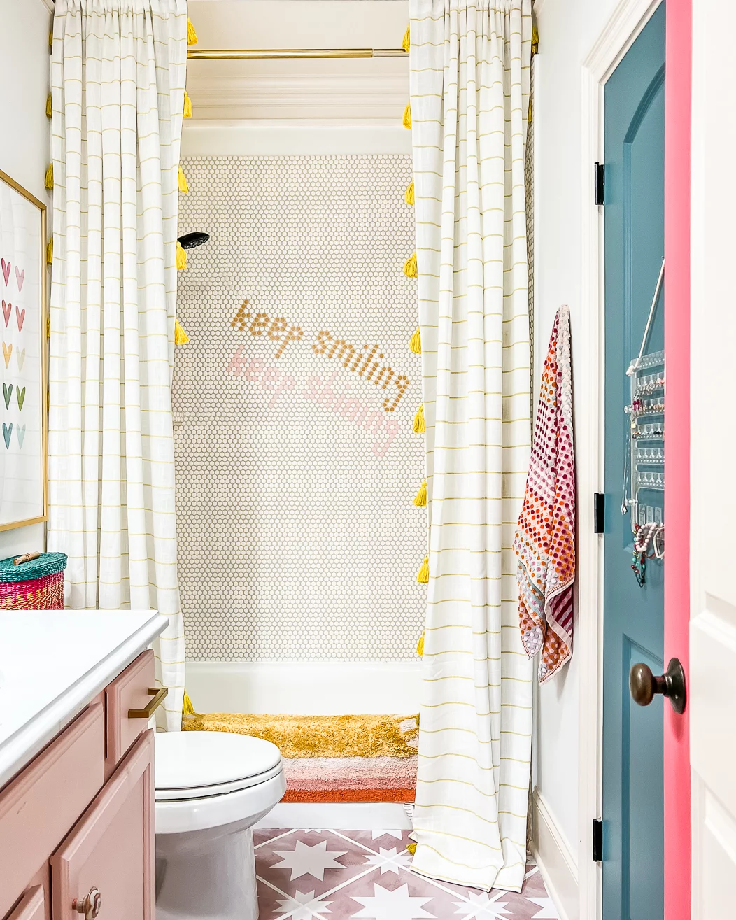 An "after" photo of a bathroom makeover on a budget features round tiles, a pink and white floor, light pink and white sink cabinet, and tub with "Keep Smiling" on the shower wall. 