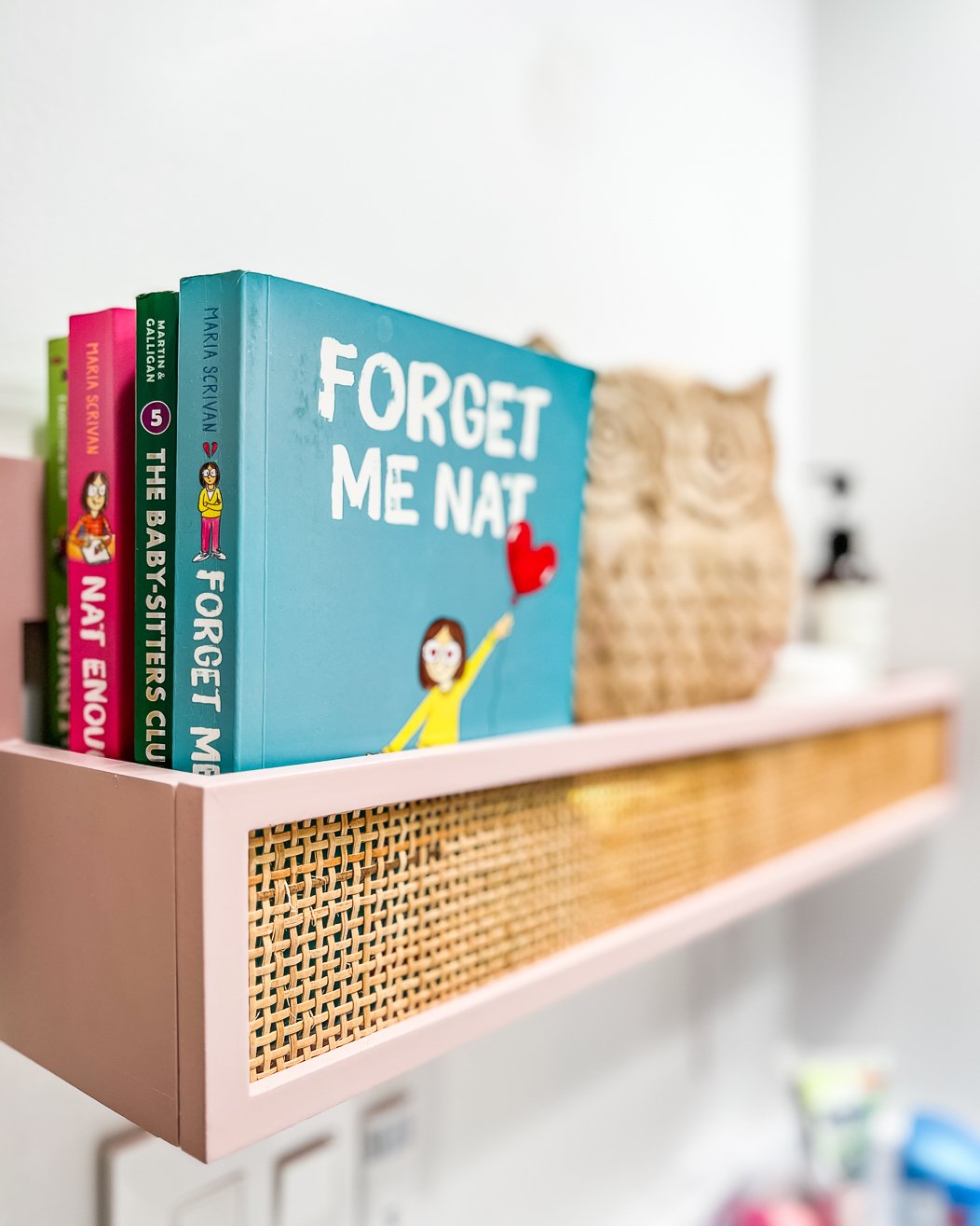 A close-up of a muted pink and caned hanging wall box full of books, and owl statue, and lotions. 