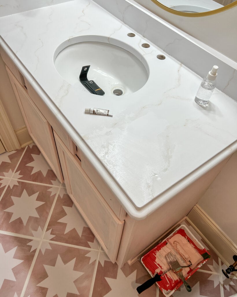 An overhead view of a bathroom countertop painted to look like white marble, with a light pink under-cabinet and geometric flooring. 